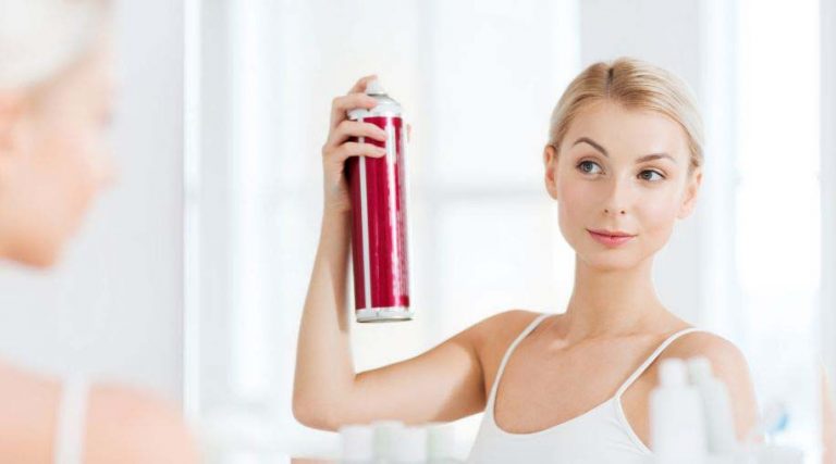 A woman standing in front of a mirror spraying hairspray into her hair.