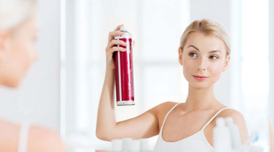A woman standing in front of a mirror spraying hairspray into her hair.