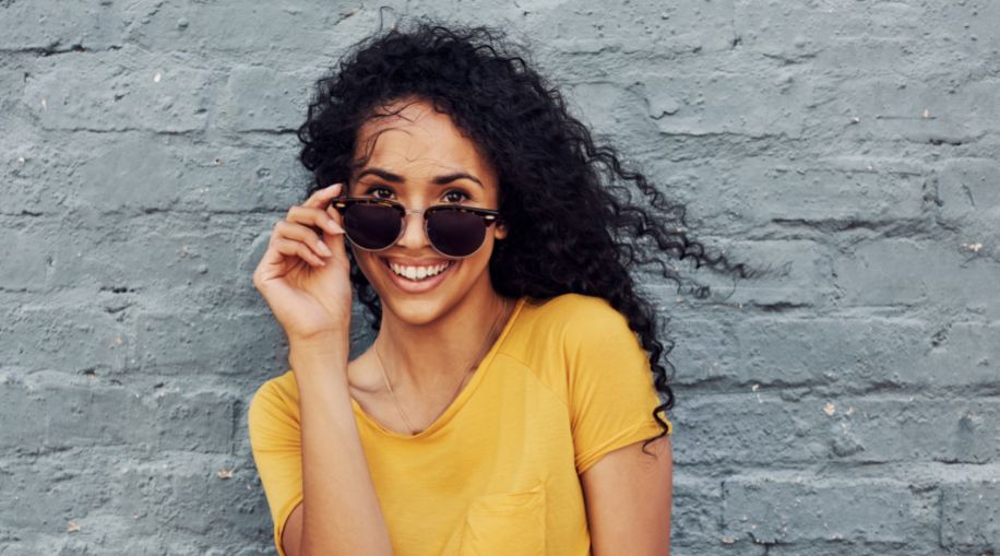 A woman in a yellow shirt holding her sunglasses down below her eyes, smiling at the camera